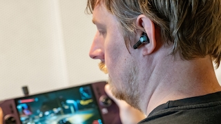Eine Person trägt einen Razer-In-Ear-Kopfhörer im Ohr. Der Ohrhörer besitzt ein glänzend schwarzes Gehäuse mit türkisfarbenem Razer-Logo auf der Außenseite. Im Hintergrund ist unscharf ein Gaming-Handheld mit laufendem Spiel zu erkennen. Die Aufnahme verdeutlicht die kompakte Bauform und den Sitz des Ohrhörers beim mobilen Spielen.