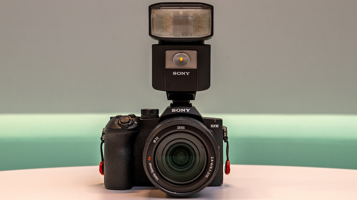 Die Sony Cbyer-shot RX10 V steht auf einem hellen Tisch. Auf der Kamera ist ein Blitzgerät montiert.