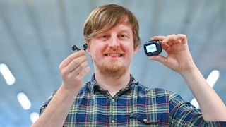 Eine Person präsentiert die JBL Live Flex 4 gemeinsam mit dem Smart Charging Case. In der einen Hand wird ein Ohrhörer gehalten, in der anderen das Ladecase mit aktivem Display. Auf dem Bildschirm des Cases ist der Eintrag „Computer“ zu sehen. Der Hintergrund ist stark unscharf und zeigt helle Lichtquellen, wodurch die Aufmerksamkeit auf Ohrhörer und Ladecase gelenkt wird. Die Aufnahme veranschaulicht die Kombination aus True-Wireless-Ohrhörern und dem multifunktionalen Ladecase mit integriertem Bildschirm.
