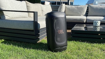 Die JBL PartyBox 130 steht auf einer Rasenfläche im Außenbereich. Im Hintergrund befindet sich eine moderne Lounge-Garnitur mit hellen Polstern sowie ein Glastisch. Der Lautsprecher besitzt ein schwarzes Gehäuse mit großflächigem Frontgitter. Hinter dem Gitter zeichnen sich die beiden Tieftöner ab. Mittig auf der Front ist ein orangefarbenes JBL-Logo angebracht. Die Aufnahme hebt die Nutzung als mobiler Outdoor-Partylautsprecher hervor.