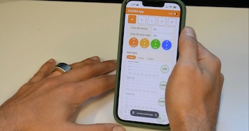 Zwei Hände einer Person liegen auf einer hellen Tischplatte. Die linke Hand trägt am Ringfinger einen breiten Smart Ring mit dunkelblauer Aussenseite und goldfarbenem Rand. Die rechte Hand hält ein Smartphone mit dunkelgrüner Hülle. Auf dem Smartphone-Display ist die CHARM App geöffnet. Oben links steht die Uhrzeit 1:27, darunter befindet sich eine orangefarbene Kopfzeile mit dem Schriftzug CHARM App und rechts ein Schalter mit der Beschriftung Light. Darunter sind fünf Schaltflächen mit den Buchstaben G, K, L, A und einem Pfeilsymbol zu sehen, wobei G orange hinterlegt ist. Es folgen zwei Eingabefelder mit den Beschriftungen Enter BG fasting und dem Wert 80 sowie Enter BG post-meal und dem Wert 130. Darunter befinden sich vier farbige Kreise mit den Angaben Glu 0 mg/dL in Orange, Ket 0 uM in Gelb, La 0 mM in Grün und Al 0 Prozent in Blau. Im Bereich Past Data sind die Reiter 2 hours, 4 hours und 8 hours zu sehen, wobei 2 hours aktiv ist. Darunter folgen drei leere Liniendiagramme mit den Beschriftungen Glucose in mg/dL, Ketone in uM und Lactate in mM, jeweils mit einem grünen Kreis mit dem Wert 0.00 daneben. Am unteren Rand ist eine Browserleiste mit der teilweise sichtbaren Adresse p-ivory.vercel.app erkennbar.