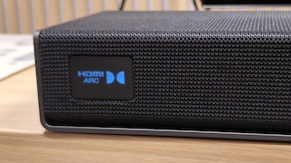Nahaufnahme der Soundbar mit Stoffbespannung. Das integrierte Display zeigt den aktuell gewählten Eingang „HDMI ARC“ sowie das Dolby-Logo an.