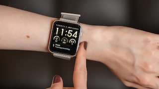 Ausgestreckter Arm mit einer Smartwatch am Handgelenk vor dunklem Hintergrund. Die Uhr hat ein eckiges Display, ein silberfarbenes Gehäuse und ein helles Textilarmband. Ein Finger der anderen Hand berührt den Rand des Displays. Auf dem schwarzen Display sind die Uhrzeit 11:54, das Datum JUN 9, der Schriftzug GARMIN, drei Anzeigen mit den Werten 64, 202 und 54 S sowie der Text Heute Trainingsvorschlag zu sehen.
