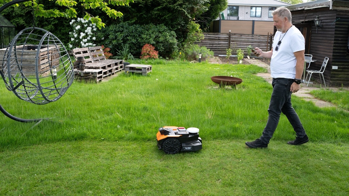 Ein Mähroboter bewegt sich über eine Rasenfläche, dahinter läuft der Tester mit Handy in der Hand. Ziel ist es, die Fläche zu kartieren.