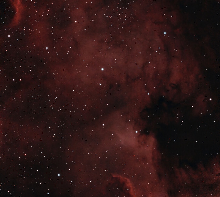 Astroaufnahme mit vielen Sternen und rötlichen Nebelstrukturen vor dunklem Himmel.