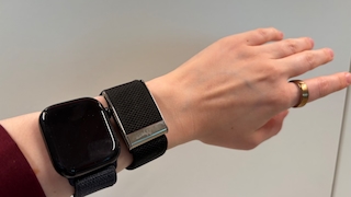 Ein ausgestreckter Arm vor einem hellgrauen Hintergrund. Am Handgelenk werden zwei Geräte nebeneinander getragen. Links befindet sich eine Smartwatch mit eckigem schwarzem Gehäuse, ausgeschaltetem dunklem Display und einem geflochtenen schwarzen Armband. Rechts daneben ist ein weiteres Gerät mit einem breiten schwarzen Textilarmband und einer silberfarbenen Metallschliesse zu sehen. Am Zeigefinger trägt die Person einen goldfarbenen Smart Ring. Die Person trägt einen dunkelroten Ärmel.
