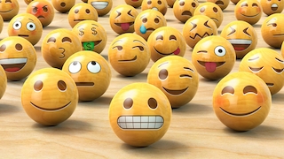 Mehrere kugelförmige Emojis mit unterschiedlichen Gesichtsausdrücken stehen dicht verteilt auf einer hellen Holzoberfläche. Zu sehen sind unter anderem lächelnde, zwinkernde, erstaunte, grinsende und herausstreckende Emojis. Einige zeigen Tränen, Geldsymbole in den Augen oder verzogene Mundformen. Die gelb-orangenen Kugeln haben eine glänzende Oberfläche mit Lichtreflexionen. Im Vordergrund befinden sich besonders große Emojis, während weitere Emojis bis in den Hintergrund reichen.