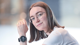 Eine Frau mit schulterlangen glatten braunen Haaren und einer runden Brille mit dünnem goldfarbenem Metallrahmen blickt in die Kamera. Sie hat den Kopf leicht zur Seite geneigt und hält die linke Hand mit angewinkelten Fingern nach oben. Sie trägt ein weisses Oberteil. Am linken Handgelenk befindet sich eine Smartwatch mit rundem Display, silberfarbenem Gehäuse und schwarzem Armband. Auf dem schwarzen Display ist ein Zifferblatt mit der Uhrzeit 11:55 in grüner Schrift und mehreren kleinen Anzeigen zu sehen. Der Hintergrund ist hell und stark unscharf mit weichen Lichtreflexen.