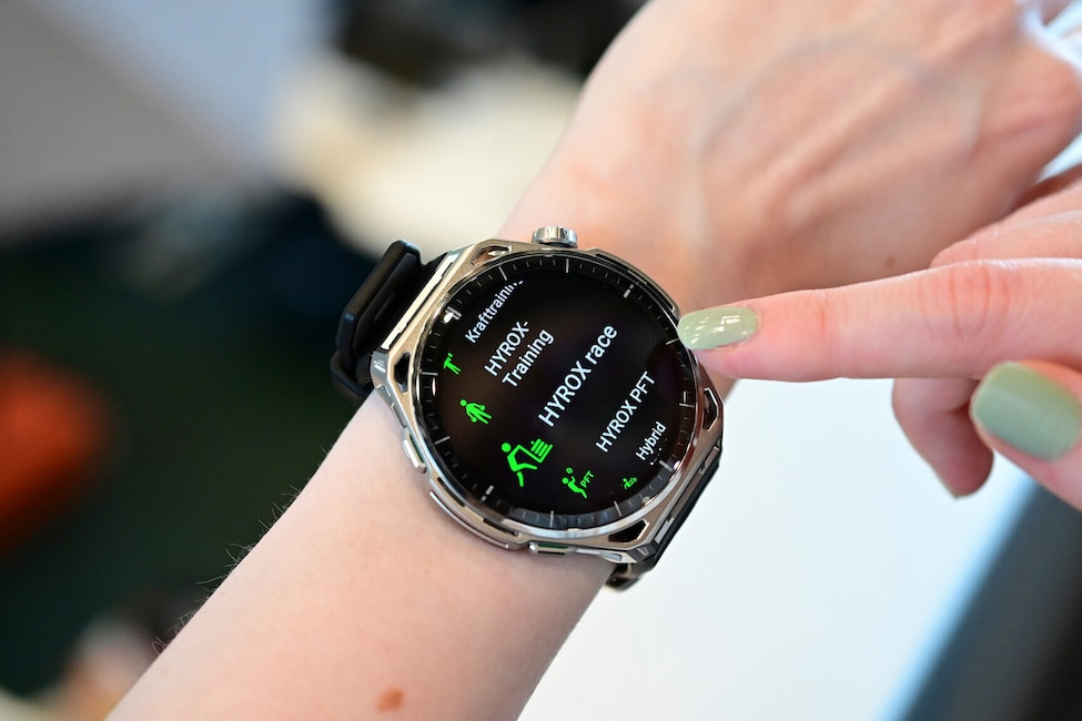 Nahaufnahme eines Handgelenks mit einer grossen Smartwatch mit rundem Display, silberfarbenem Gehäuse mit auffälliger Lünette und schwarzem Silikonarmband. Ein Finger der anderen Hand mit hellgrün lackiertem Nagel berührt den rechten Rand des Displays. Auf dem schwarzen Display ist eine Sportarten-Auswahl mit grünen Icons und weisser Schrift zu sehen. Aufgelistet sind von oben nach unten Krafttraining, HYROX-Training, HYROX race, HYROX PFT und Hybrid. An der rechten Gehäuseseite befinden sich eine Krone und Tasten. Der Hintergrund ist unscharf und zeigt einen hellen Innenraum.