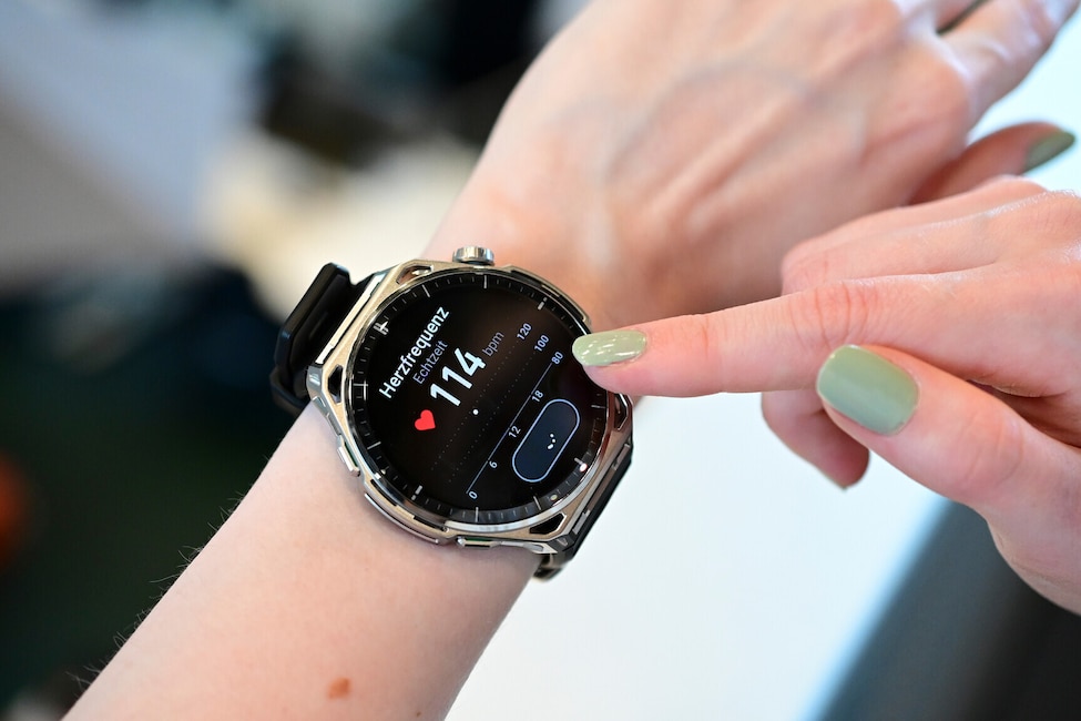 Nahaufnahme eines Handgelenks mit einer grossen Smartwatch mit rundem Display, silberfarbenem Gehäuse mit auffälliger Lünette und schwarzem Silikonarmband. Ein Finger der anderen Hand mit hellgrün lackiertem Nagel berührt den rechten Rand des Displays. Auf dem schwarzen Display steht oben die Überschrift Herzfrequenz und darunter Echtzeit. In der Mitte ist ein rotes Herzsymbol mit der grossen Zahl 114 und der Einheit bpm zu sehen. Rechts befindet sich eine senkrechte Skala mit den Werten 80, 100 und 120. Darunter ist ein Diagrammbereich mit einer Zeitachse von 0 über 6, 12 und 18 sowie einer ovalen Schaltfläche erkennbar. An der rechten Gehäuseseite befinden sich eine Krone und Tasten. Der Hintergrund ist unscharf und zeigt einen hellen Innenraum.