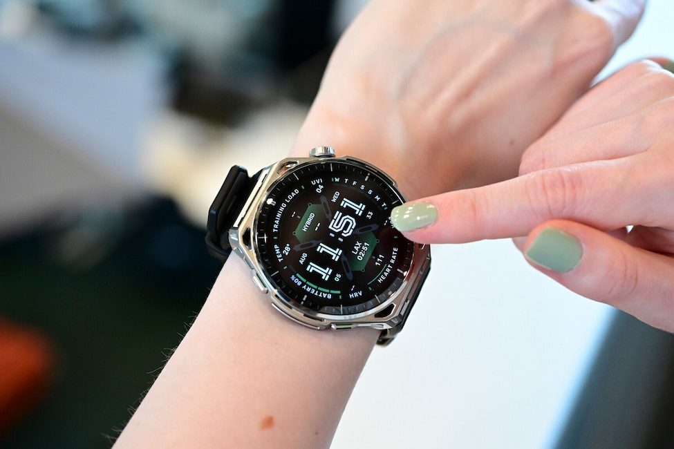 Nahaufnahme eines Handgelenks mit einer grossen Smartwatch mit rundem Display, silberfarbenem Gehäuse mit auffälliger Lünette und schwarzem Silikonarmband. Ein Finger der anderen Hand mit hellgrün lackiertem Nagel berührt den rechten Rand des Displays. Auf dem schwarzen Display ist ein Zifferblatt mit der grossen Uhrzeit 11:51 in weisser Schrift zu sehen. Rundherum sind mehrere kleine Anzeigen angeordnet: oben TRAINING LOAD, UVI 04 und WED, links TEMP 28 Grad und AUG 05, rechts 33, LAX 02:51 und HEART RATE 111 sowie unten BATTERY 80 Prozent und HRV. An der rechten Gehäuseseite befinden sich eine Krone und Tasten. Der Hintergrund ist unscharf und zeigt einen hellen Innenraum.