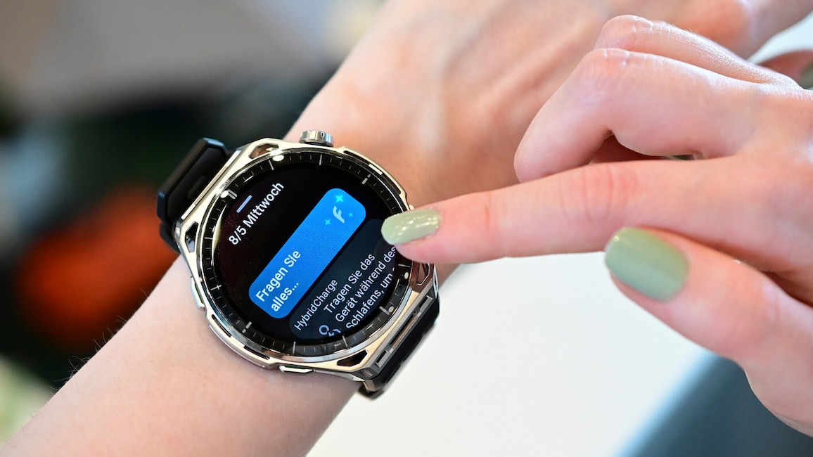 Nahaufnahme eines Handgelenks mit einer grossen Smartwatch mit rundem Display, silberfarbenem Gehäuse mit auffälliger Lünette und schwarzem Silikonarmband. Ein Finger der anderen Hand mit hellgrün lackiertem Nagel berührt den rechten Rand des Displays. Auf dem schwarzen Display steht oben links das Datum 8/5 Mittwoch. Rechts daneben befindet sich eine blaue Kachel mit dem Text Fragen Sie alles und einem Sternchen-Symbol. Darunter ist eine weitere Kachel mit der Überschrift HybridCharge und dem teilweise sichtbaren Text Tragen Sie das Gerät während des Schlafens, um zu sehen. An der rechten Gehäuseseite befinden sich eine Krone und Tasten. Der Hintergrund ist unscharf und zeigt einen hellen Innenraum.