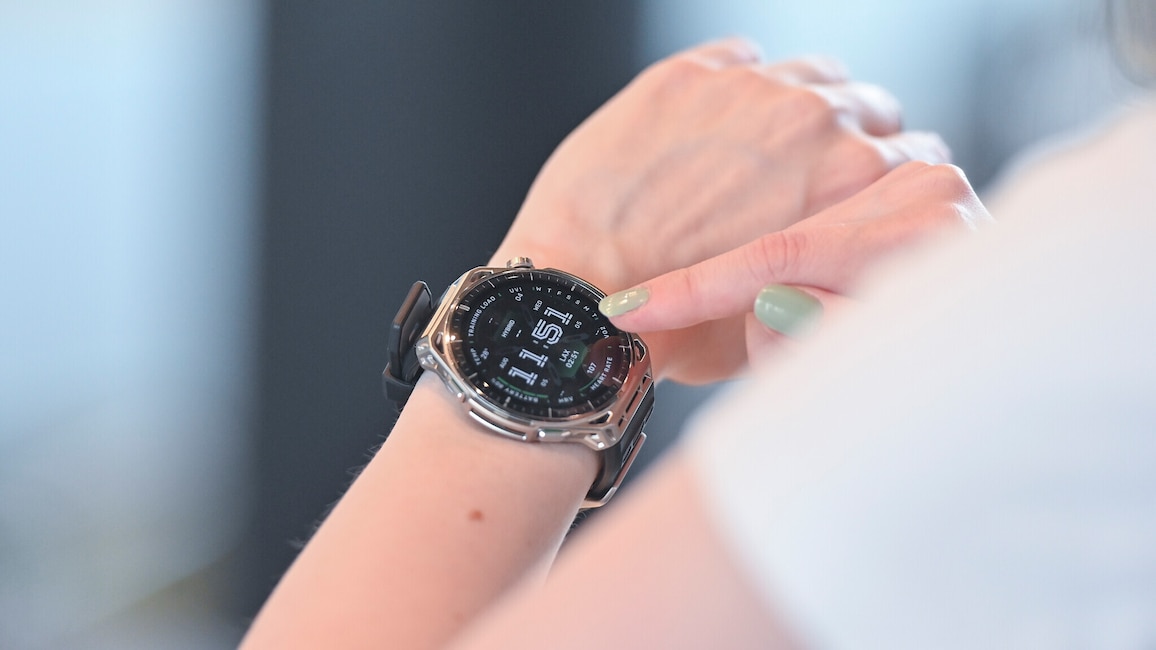 Nahaufnahme eines Handgelenks mit einer grossen Smartwatch mit rundem Display, silberfarbenem Gehäuse mit auffälliger Lünette und schwarzem Silikonarmband. Ein Finger der anderen Hand mit hellgrün lackiertem Nagel nähert sich dem rechten Rand des Displays. Auf dem schwarzen Display ist ein Zifferblatt mit der grossen Uhrzeit 11:51 in weisser Schrift zu sehen. Rundherum sind mehrere kleine Anzeigen angeordnet, darunter TRAINING LOAD, UVI 04, WED, TEMP, AUG, HYBRID, LAX 02:51, 107, HEART RATE, VO2, BATTERY und HRV. An der linken Gehäuseseite ist eine Krone erkennbar. Der Hintergrund ist unscharf und in Blau- und Grautönen gehalten.