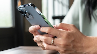 Nahaufnahme von zwei Händen, die ein Smartphone mit silberfarbenem Gehäuse halten. Auf der Rückseite des Geräts sind drei große Kameralinsen, ein Blitz und mehrere seitliche Tasten sichtbar. Eine Hand trägt einen Ring am Finger. Der Hintergrund ist unscharf und zeigt helle Fensterflächen sowie Teile eines Innenraums.