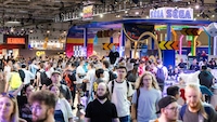 Eine Messehalle mit dichtem Besucheraufkommen ist aus erhöhter Perspektive fotografiert. Mehrere groĂe MessestĂ€nde mit auffĂ€lliger Beleuchtung dominieren die Szene. Besonders sichtbar sind die Markenauftritte von SEGA und Sonic Racing CrossWorlds. ZusĂ€tzlich sind weitere Standbeschriftungen wie âREANIMALâ erkennbar. Zahlreiche Besucher bewegen sich zwischen den AusstellungsflĂ€chen und Teststationen.