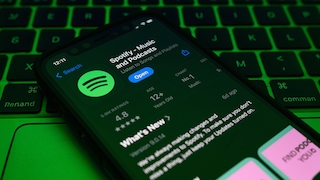 Nahaufnahme eines Smartphones, das schräg auf einer Tastatur mit grüner Hintergrundbeleuchtung liegt. Auf dem Display ist die App-Seite von „Spotify – Music and Podcasts“ im App Store zu sehen. Sichtbar sind das Spotify-Logo, ein blauer „Open“-Button, eine Bewertung von 4,8 Sternen, die Altersfreigabe „12+ Years Old“ sowie der Abschnitt „What’s New“. Die Tastatur füllt den Hintergrund und ist durch die grüne Beleuchtung deutlich hervorgehoben.