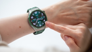 Nahaufnahme eines Handgelenks mit einer grossen Smartwatch mit rundem Display, silberfarbenem Gehäuse und grünem Silikonarmband vor einem hellen Hintergrund. Ein Finger der anderen Hand berührt den rechten Rand des Displays. Auf dem grünen Zifferblatt sind grosse weisse Ziffern von 1 bis 12, weisse Zeiger und ein oranger Sekundenzeiger zu sehen. Oben mittig steht das Datum Mi. 26. Links ist eine kleine Anzeige mit dem Wert 111 und einem Herzsymbol, rechts eine mit dem Wert 3506 und einem Schrittsymbol. Unten mittig ist eine Wetteranzeige mit einem Wolkensymbol, der Temperatur 23 Grad und den Werten 25 Grad und 14 Grad zu sehen. An der rechten Gehäuseseite befindet sich eine Krone.
