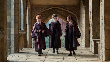 Drei Kinder in dunklen Hogwarts-Umhängen gehen durch einen steinernen Gang mit Bögen und Säulen. Im Hintergrund ist ein weiterer Torbogen zu sehen, durch den Tageslicht einfällt.