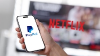 Eine Hand hält ein Smartphone mit dem PayPal-Logo auf dem Display. Im unscharfen Hintergrund sind ein Bildschirm mit dem Netflix.