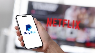 Eine Hand hält ein Smartphone mit dem PayPal-Logo auf dem Display. Im unscharfen Hintergrund sind ein Bildschirm mit dem Netflix.