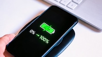 Smartphone auf einem kabellosen Ladepad. Auf dem Display ist ein grünes Batteriesymbol mit Blitz zu sehen, darunter die Anzeige „0 % → 100 %“. Im Hintergrund ist unscharf eine weiße Tastatur zu erkennen. Die Aufnahme vermittelt das schnelle Aufladen eines Akkus.