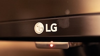 Nahaufnahme eines LG-Fernsehers. Auf dem schwarzen Rahmen des Geräts ist das weiße LG-Logo zu sehen. Unterhalb des Fernsehers leuchtet eine kleine rote Status-LED, der Hintergrund ist unscharf und warm beleuchtet.