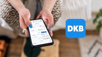 Eine Person hält ein Smartphone mit geöffneter DKB-Banking-App in beiden Händen. Auf dem Display sind Kontostand und mehrere Kontobewegungen zu sehen. Rechts daneben ist das blaue DKB-Logo eingeblendet. Im Hintergrund sind unscharf ein Wohnraum mit Holzboden und eine Zimmerpflanze zu erkennen.