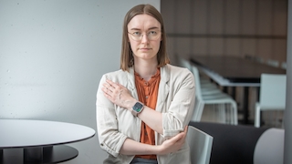 Eine Frau mit schulterlangen glatten braunen Haaren und einer runden Brille mit dünnem Metallrahmen steht mit vor dem Körper verschränkten Armen und blickt in die Kamera. Sie trägt eine rostbraune Bluse unter einem hellbeigen Blazer. Am linken Handgelenk befindet sich eine Smartwatch mit eckigem Display, hellem Gehäuse und beigem Armband. Auf dem Display sind der Wert 79 und ein grünes Symbol zu erkennen. Der Hintergrund zeigt einen hellen Innenraum mit einem runden Tisch, mehreren Stühlen und einer grauen Wand.