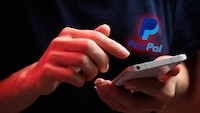 Nahaufnahme von zwei Händen, die ein weißes Smartphone halten und bedienen. Über dem Smartphone ist das blau-türkise PayPal-Logo mit dem Schriftzug „PayPal“ eingeblendet. Die Szene ist in dunkler Umgebung aufgenommen, mit roter und blauer Beleuchtung auf den Händen und im Hintergrund.