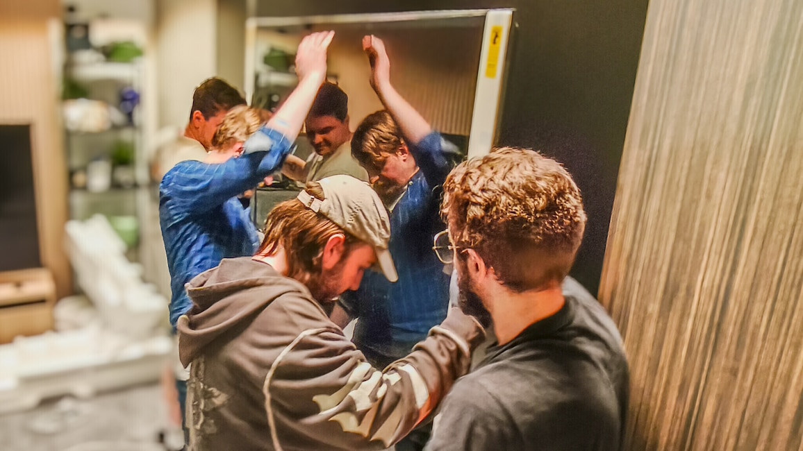 Ein Team von vier Männern bringt einen großen Flachbild-TV in ein Zimmer. Sie wirken angestrengt und konzentriert.