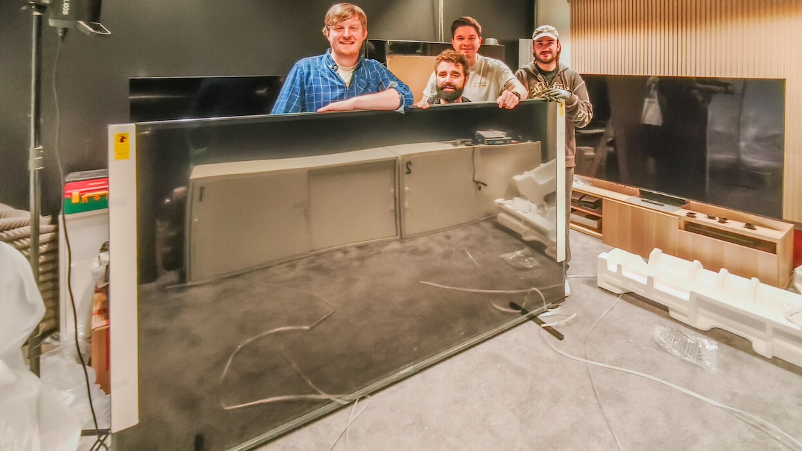 Ein Team von vier Männern steht glücklich hinter einem großen Fernseher, der mitten in einem Zimmer auf dem Steinboden steht. Der TV-Bildschirm spiegelt eine weiße Anrichte mit vier Türen. Davor liegen Verpackungsbänder. Rechts liegt Verpackungs-Styropor vor einem Sideboard, auf dem ein weiterer TV steht.