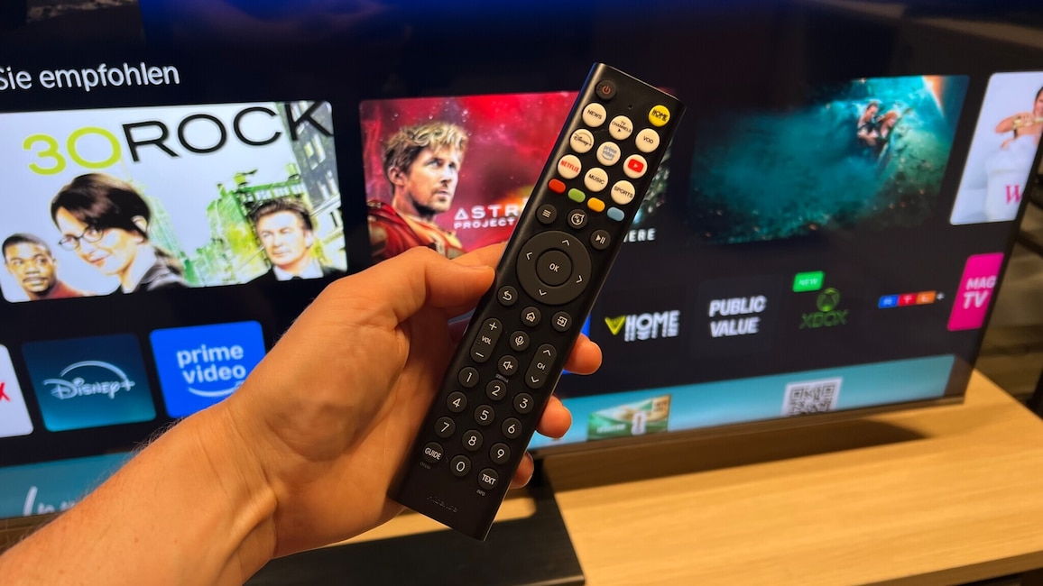 Eine Hand hält die Fernbedienung des Hisense 100U7DSE, im Hintergrund steht ein Fernseher.