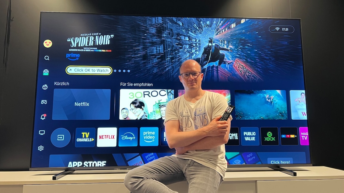 Redakteur Dennis Ellenberger sitzt entspannt, Fernbedienung in der Hand, auf einem beigen Sideboard vor einem dort aufgestellten großen Fernseher. Auf dem TV-Bildschirm sind Kacheln von Streamingiensten wie Netflix und Prime Video zu sehen, außerdem ein Vorschaubild der US-Serie "Spider Noir" und darunter ein Button "Click OK to watch".