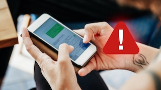 Nahaufnahme von zwei Händen, die ein Smartphone in einer aufklappbaren Hülle halten. Auf dem Display ist eine geöffnete Nachrichtenanwendung mit einer grünen Sprechblase und einer eingeblendeten Tastatur zu sehen. Rechts neben dem Smartphone ist ein großes rotes Warnsymbol mit einem weißen Ausrufezeichen platziert. Im Hintergrund sind unscharfe Bereiche eines Innenraums sowie die Kante eines Tisches zu erkennen. An einem Handgelenk ist eine sichtbare Tätowierung zu sehen.