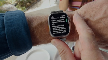 Nahaufnahme einer Apple Watch Ultra mit silbernem Milanaise-Armband am Handgelenk einer Person. Auf dem Display steht oben rechts Rewind und die Uhrzeit 10:09, darunter der transkribierte Text Todays specials are the Heirloom Tomato BLT sowie eine Aufzählung von Gerichten wie Caesar salad, cobb salad, lentil soup, chicken Alfredo, smoked salmon und avocado tartine. Rechts am Displayrand ein grüner Balken. Im unscharfen Hintergrund eine sonnige Terrasse.