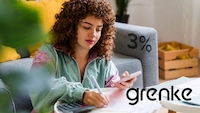 Eine Frau sitzt vor einem Tisch und schreibt auf Unterlagen. Sie hält ein Smartphone in der Hand, daneben liegt ein Taschenrechner. Rechts im Bild sind der Schriftzug „grenke“ und „3%“ eingeblendet. Im Hintergrund sind ein Sofa, Kissen und eine Holzkiste zu sehen.