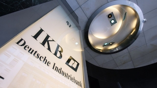 Ein Schild mit der Aufschrift „IKB Deutsche Industriebank“ ist an einer Gebäudefassade zu sehen. Rechts daneben befindet sich ein runder, beleuchteter Gebäudeteil mit kleinen Leuchten.