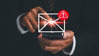 Nahaufnahme einer Person in dunkler Geschäftskleidung, die ein Smartphone in der Hand hält und mit einem Finger auf ein eingeblendetes Symbol eines Briefumschlags tippt. Über dem Umschlagsymbol befindet sich ein rotes Warnsymbol mit Ausrufezeichen. Das Motiv ist vor dunklem Hintergrund aufgenommen, der Bereich um den Berührungspunkt des Fingers ist mit einem hellen Lichteffekt hervorgehoben.