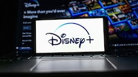 Ein Smartphone mit dem Disney+-Logo auf dem Display liegt auf der Tastatur eines Laptops. Im Hintergrund ist unscharf die Startseite von Disney+ mit verschiedenen Serien- und Filmkacheln zu sehen. Die Szene ist dunkel beleuchtet, das helle Display steht im Mittelpunkt des Bildes.
