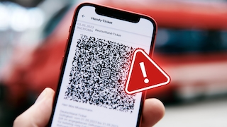 Nahaufnahme eines Smartphones mit digitalem Deutschlandticket und QR-Code auf dem Display. Vor dem Bildschirm ist ein rotes Warnsymbol mit Ausrufezeichen eingeblendet. Im Hintergrund ist unscharf ein rotes Fahrzeug zu sehen.