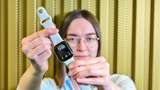 Eine Frau mit schulterlangen glatten braunen Haaren und einer runden Brille mit dünnem Metallrahmen hält mit beiden Händen eine Smartwatch in die Kamera. Die Uhr ist scharf im Vordergrund, ihr Gesicht unscharf im Hintergrund. Sie trägt eine hellblaue Bluse. Die Smartwatch hat ein eckiges Display, ein silberfarbenes Gehäuse und ein weisses Silikonarmband, das nach oben und unten gehalten wird. Auf dem schwarzen Display sind das Datum FR. 18. SEPT., die Uhrzeit 13:52 sowie drei kleine Icons in Grün, Weiss und Rot zu sehen. Der Hintergrund zeigt eine gelb-goldfarbene Wand mit senkrechter Struktur.