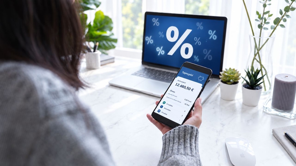Eine Person hält ein Smartphone mit einer Tagesgeld-Ansicht in der Hand. Auf dem Display sind Kontostand, Zinsen und Menüeinträge zu sehen. Im Hintergrund steht ein Laptop mit großem Prozentzeichen auf dem Bildschirm. Auf dem Tisch stehen mehrere kleine Pflanzen, eine Kerze und eine Computermaus.