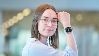 Eine Frau mit schulterlangen glatten braunen Haaren und einer runden Brille mit dünnem goldfarbenem Metallrahmen blickt in die Kamera. Sie ist seitlich zur Kamera gedreht und hat den angewinkelten rechten Arm an ihren Kopf gelegt, die Hand zur Faust geballt. Sie trägt eine hellblaue Bluse. Am rechten Handgelenk befindet sich eine grosse Smartwatch mit eckigem Display, titanfarbenem Gehäuse und einem beigefarbenen Armband. Auf dem Display ist die Uhrzeit 14:04 in einem runden Zifferblatt mit grünen und weissen Elementen sowie einem Plus-Symbol zu sehen. Der Hintergrund ist stark unscharf und zeigt einen hellen Innenraum mit runden Lichtreflexen.