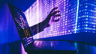 Geöffneter Laptop auf einer dunklen Oberfläche vor einem blau-violett beleuchteten Hintergrund mit zahlreichen Zeichen und Zahlen. Auf dem Laptop-Bildschirm ist ein stilisiertes Vorhängeschloss zu sehen. Aus dem Bereich des Bildschirms ragt eine ausgestreckte Hand in den Raum. Das Bild ist in starken Blau- und Violetttönen gehalten und zeigt eine digitale, technisch geprägte Szene.