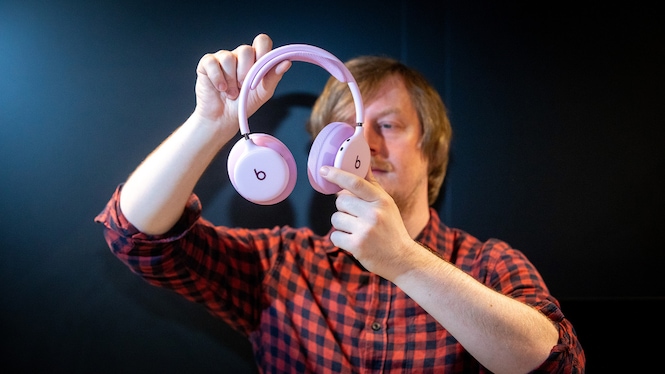 Eine Person hält den rosafarbenen Beats-Over-Ear-Kopfhörer mit beiden Händen vor die Kamera. Der Kopfhörer ist vollständig aufgeklappt und frontal ausgerichtet. Gut sichtbar sind die großen runden Hörmuscheln, die Stoffpolster, der schlanke Kopfbügel sowie die Beats-Logos auf beiden Außenseiten. Der dunkle Hintergrund und die gerichtete Beleuchtung setzen den hellen Kopfhörer deutlich vom Umfeld ab.