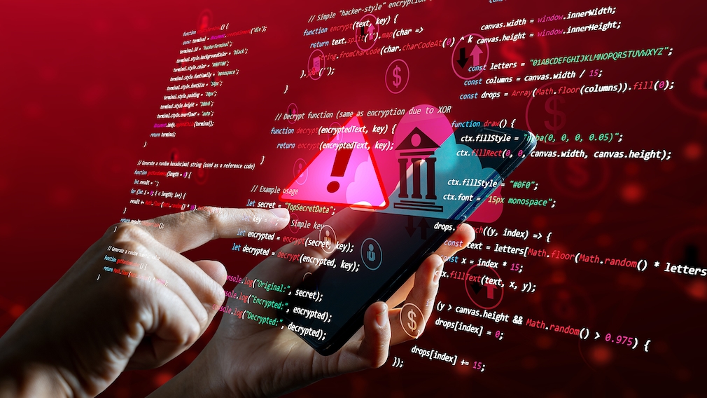 Eine Person hält ein Smartphone in einer Hand und tippt mit einem Finger der anderen Hand auf den Bildschirm. Über dem Smartphone sind grafische Symbole eingeblendet, darunter ein Warndreieck mit Ausrufezeichen, ein Bankgebäude, Pfeile sowie Währungssymbole. Im Hintergrund ist ein roter Farbverlauf mit unscharfen Lichteffekten zu sehen. Über das gesamte Bild legen sich zahlreiche Zeilen mit Programmcode in weißer, grüner und roter Schrift.