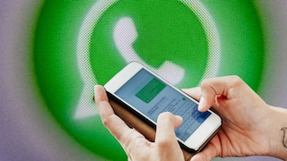 Zwei Hände halten ein Smartphone im Querformat. Auf dem Bildschirm des Smartphones ist eine geöffnete Chat-Anwendung mit einer Bildschirmtastatur zu sehen. Im Hintergrund befindet sich stark vergrößert ein grünes WhatsApp-Logo auf einer pixeligen Bildschirmdarstellung. Das Smartphone und die Hände stehen im Vordergrund, während das Logo unscharf im Hintergrund erscheint. Die Bildgestaltung wird von Grün-, Weiß- und Grautönen geprägt.
