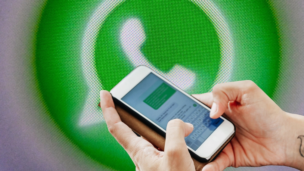 Zwei Hände halten ein Smartphone im Querformat. Auf dem Bildschirm des Smartphones ist eine geöffnete Chat-Anwendung mit einer Bildschirmtastatur zu sehen. Im Hintergrund befindet sich stark vergrößert ein grünes WhatsApp-Logo auf einer pixeligen Bildschirmdarstellung. Das Smartphone und die Hände stehen im Vordergrund, während das Logo unscharf im Hintergrund erscheint. Die Bildgestaltung wird von Grün-, Weiß- und Grautönen geprägt.
