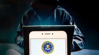 Unscharf dargestellte Person mit Kapuze sitzt hinter einem aufgeklappten Laptop in einer dunklen Umgebung. Im Vordergrund steht ein Smartphone, dessen Bildschirm ein rundes Emblem des Federal Bureau of Investigation (FBI) zeigt. Das Smartphone wird von hellem Licht angestrahlt und dominiert das Bild, während die Person und der Laptop im Hintergrund dunkel und unscharf bleiben. Die Szene ist überwiegend in dunklen Blau- und Schwarztönen gehalten.