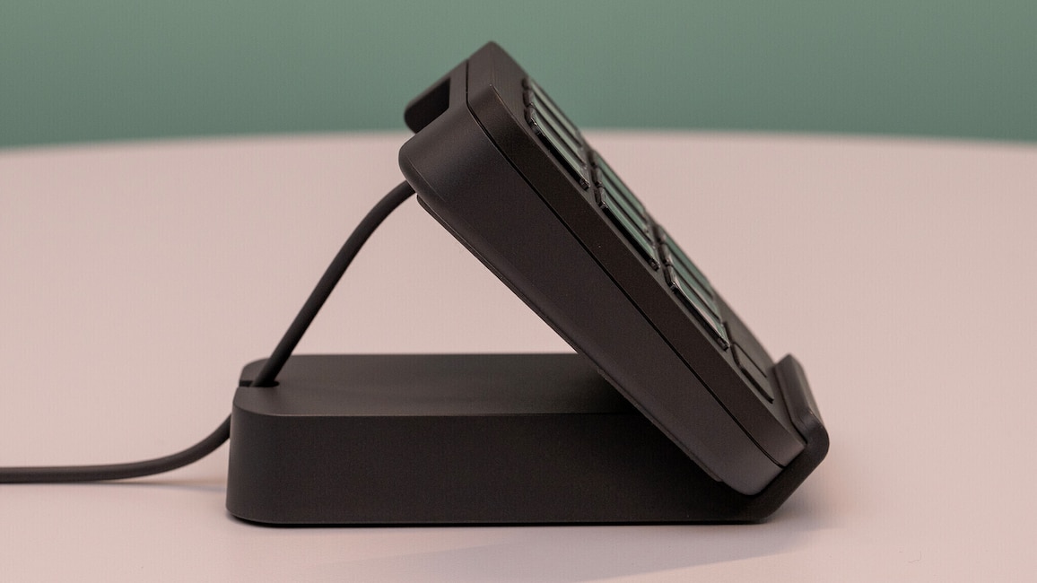 Das Logitech MX Keypad steht in der mitgelieferten Halterung auf einem weißen Tisch vor einem grünen Hintergrund.